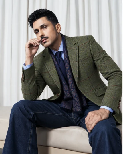 Man wearing a men's dark olive tweed jacket with rust and navy checked detailing, styled with a blue shirt, navy tie, patterned scarf, and dark denim jeans.